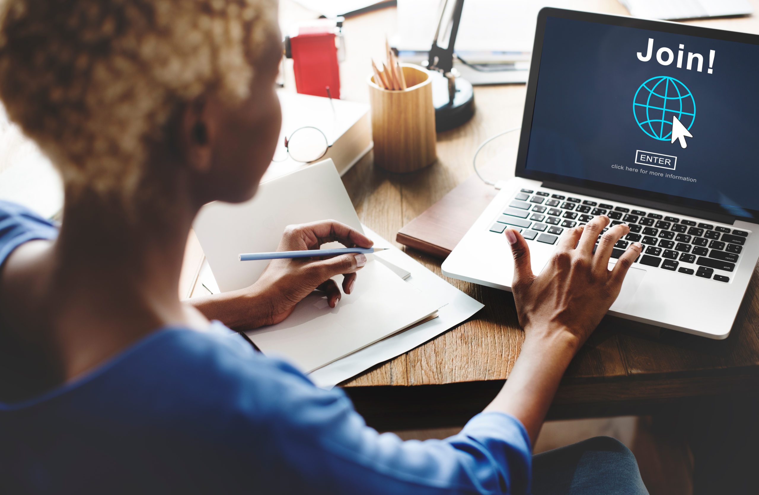 Woman sits at table, looking away from camera at a laptop with one hand on the keyboard and the other holding a pen to take notes; the laptop screen says "join"