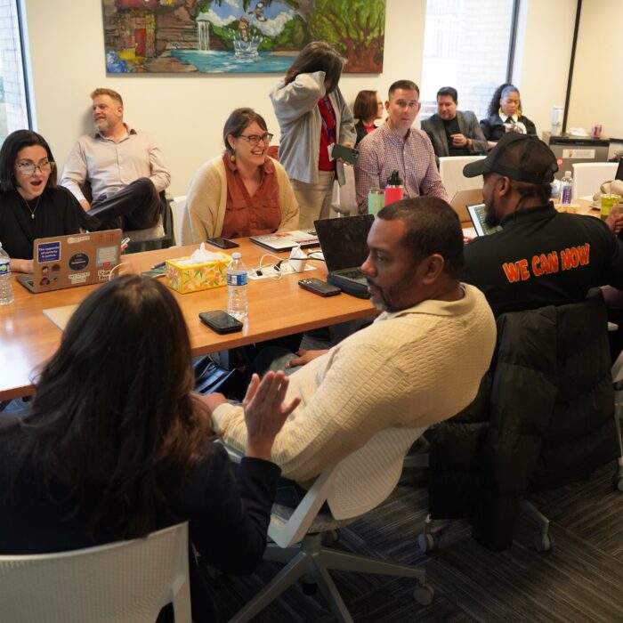 A group of people sit around a conference room table talking with water bottles and papers on the table