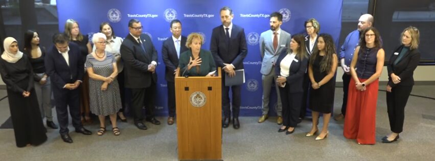 A group of people, including county commissioners, gather around a podium as a woman speaks